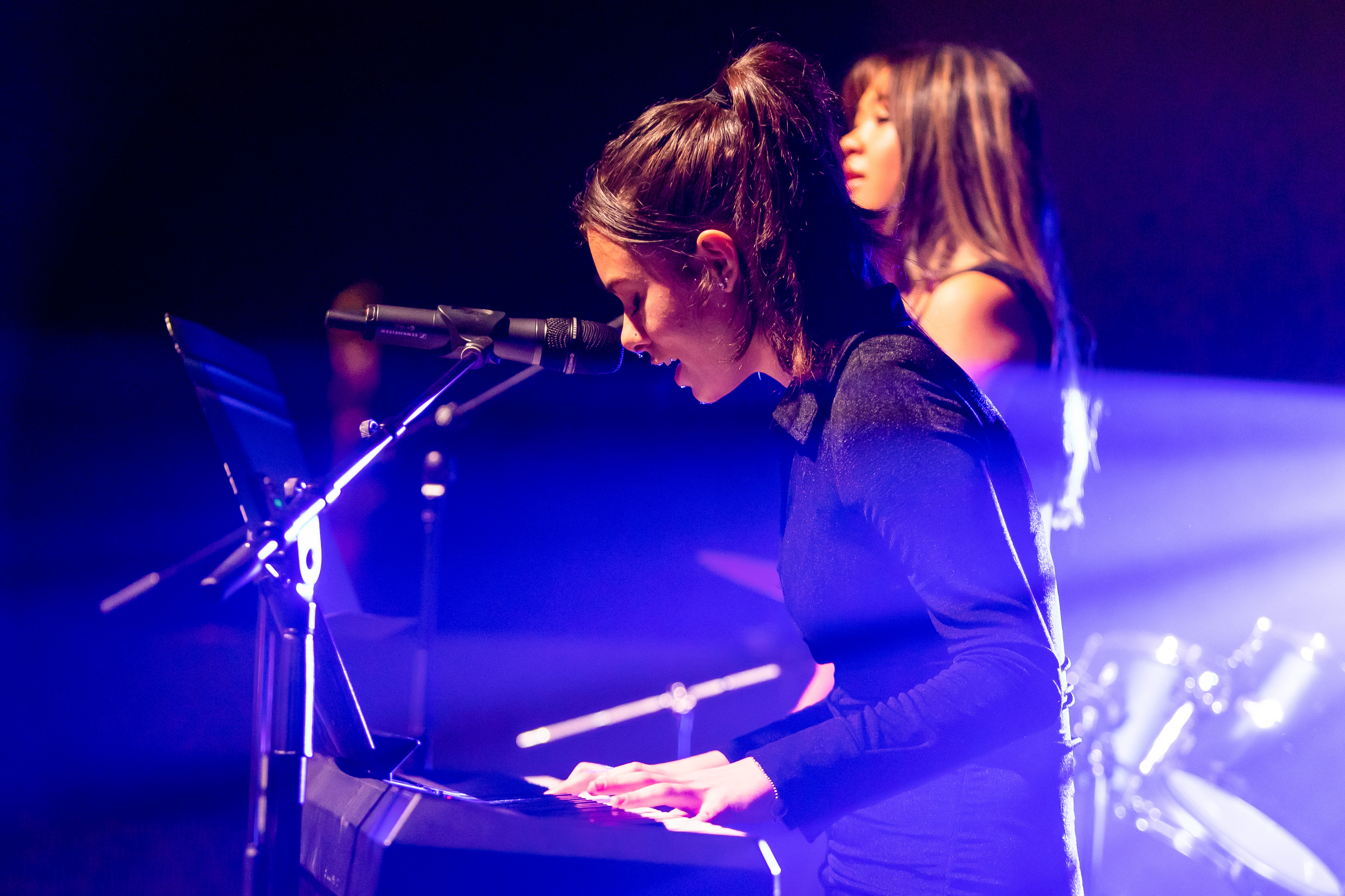 Student singing while playing keyboard on stage with microphone and band equipment under coloured lighting
