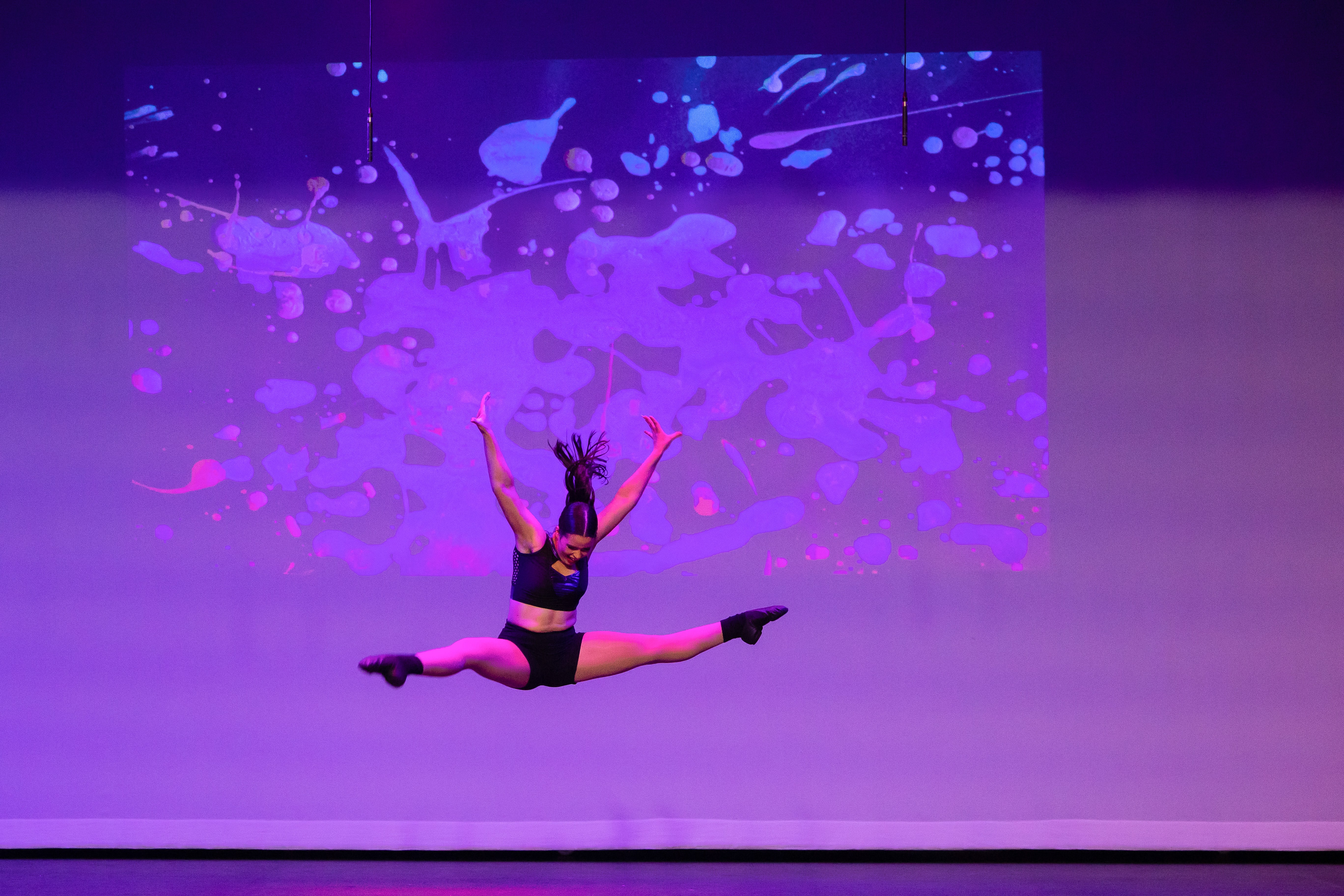 Student leaping on stage with arms raised during a contemporary dance performance with abstract projection backdrop