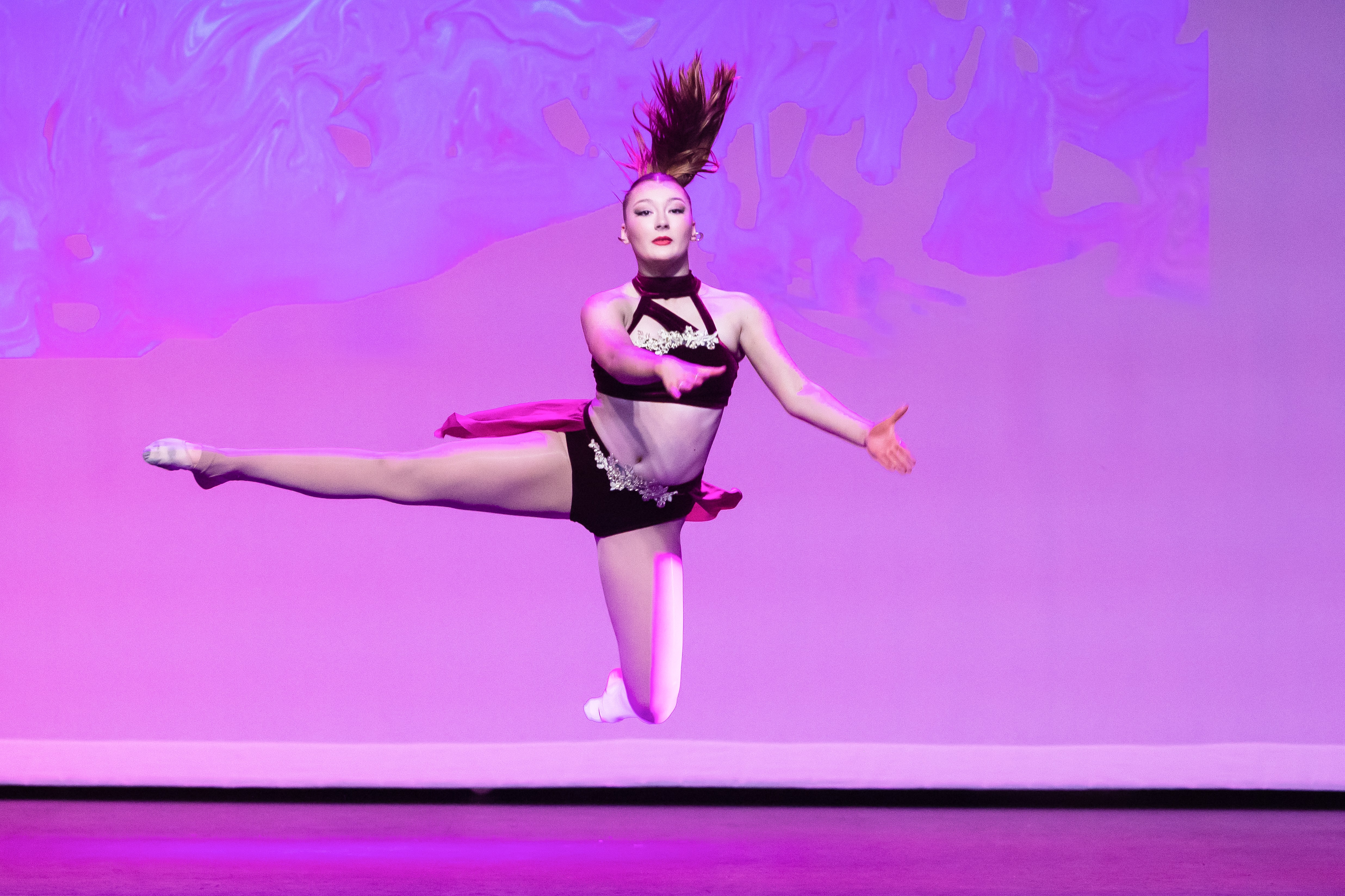 Student performing a high jump split during a contemporary dance solo under purple stage lighting