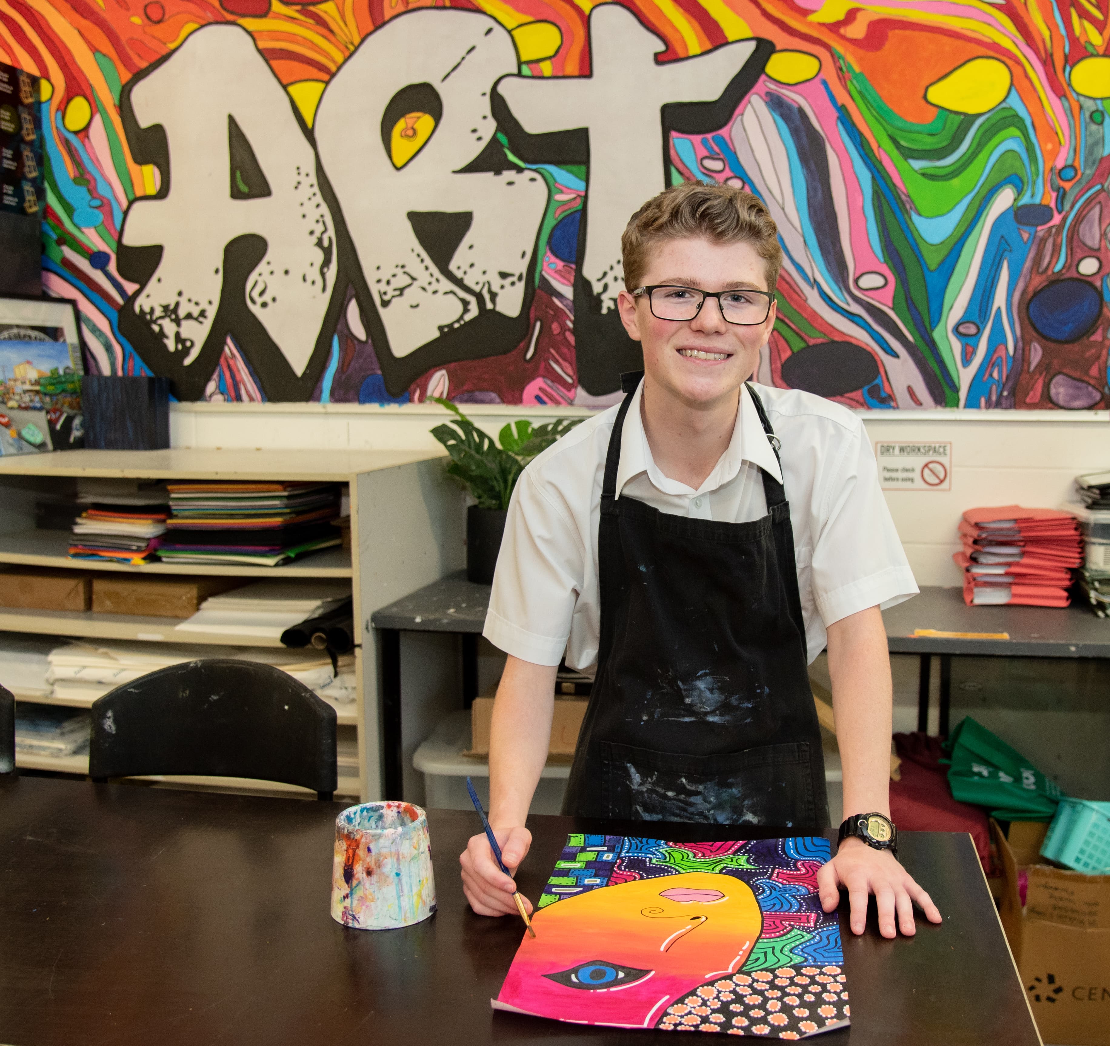 Student painting a vibrant artwork in an art classroom with supplies and projects visible in the background