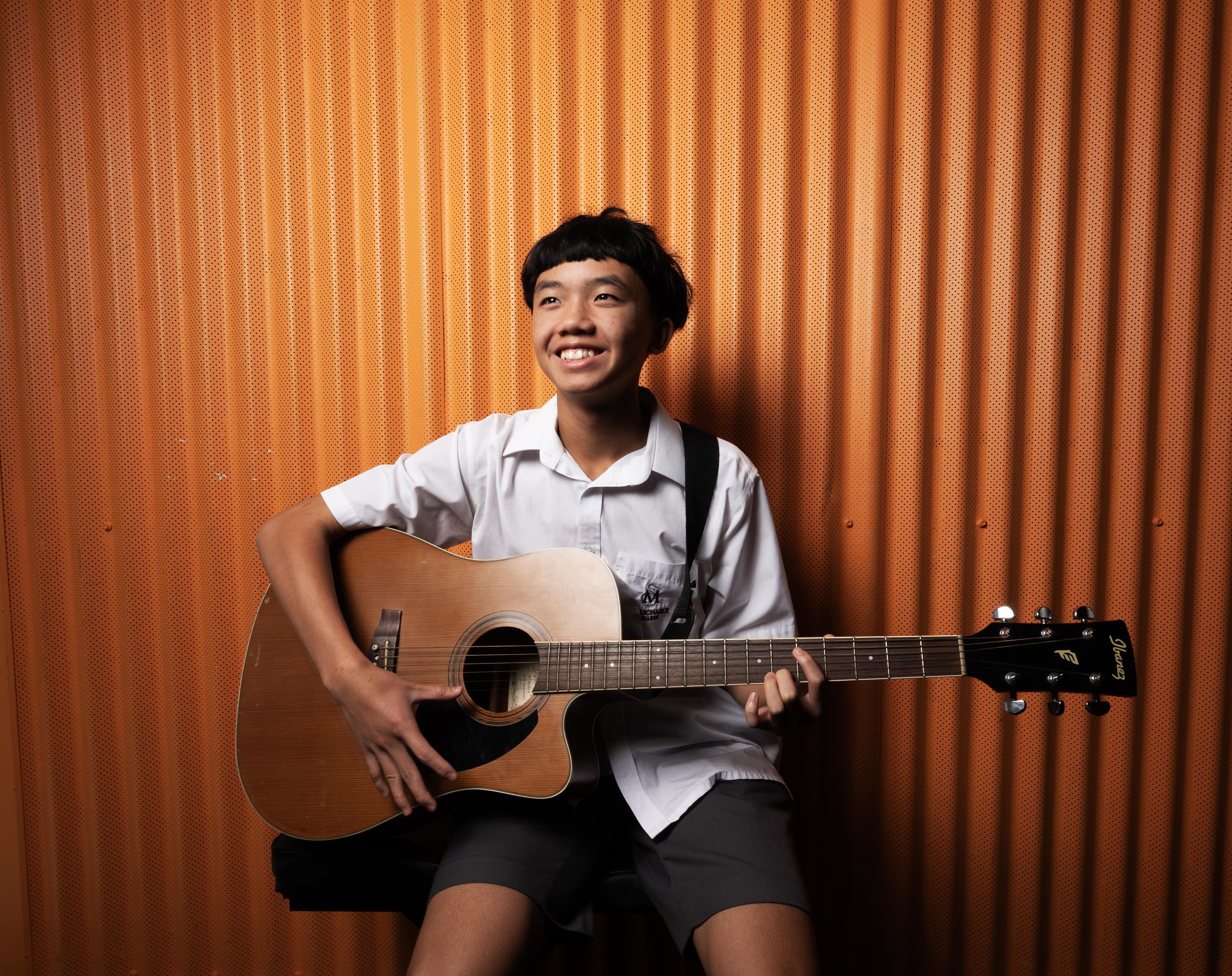 Student playing acoustic guitar indoors against textured orange backdrop