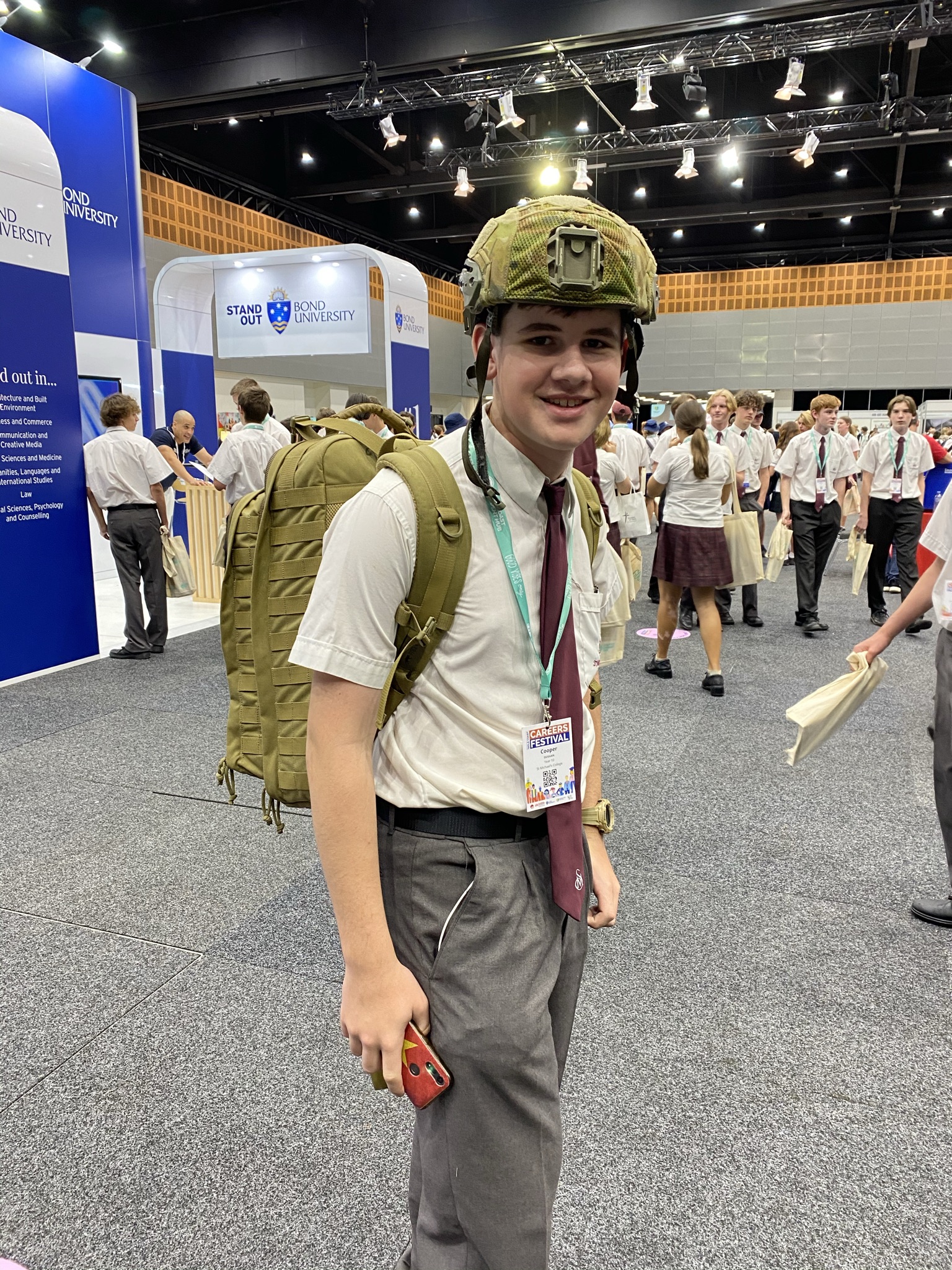 Student wearing school uniform and protective helmet with backpack at a careers expo, standing among exhibition booths and other students