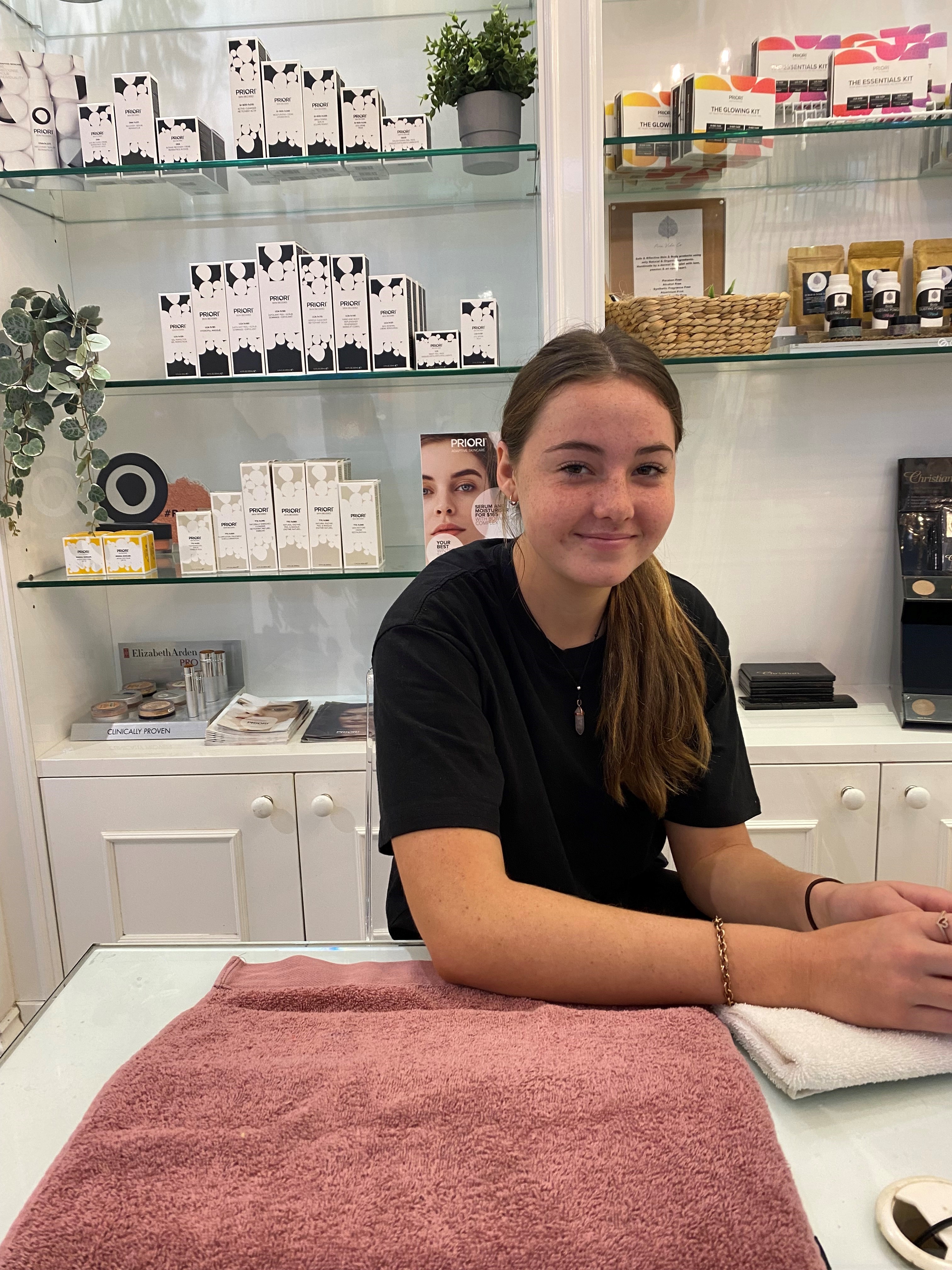 Student working in a beauty therapy setting, seated at a treatment station with skincare products displayed on shelves behind