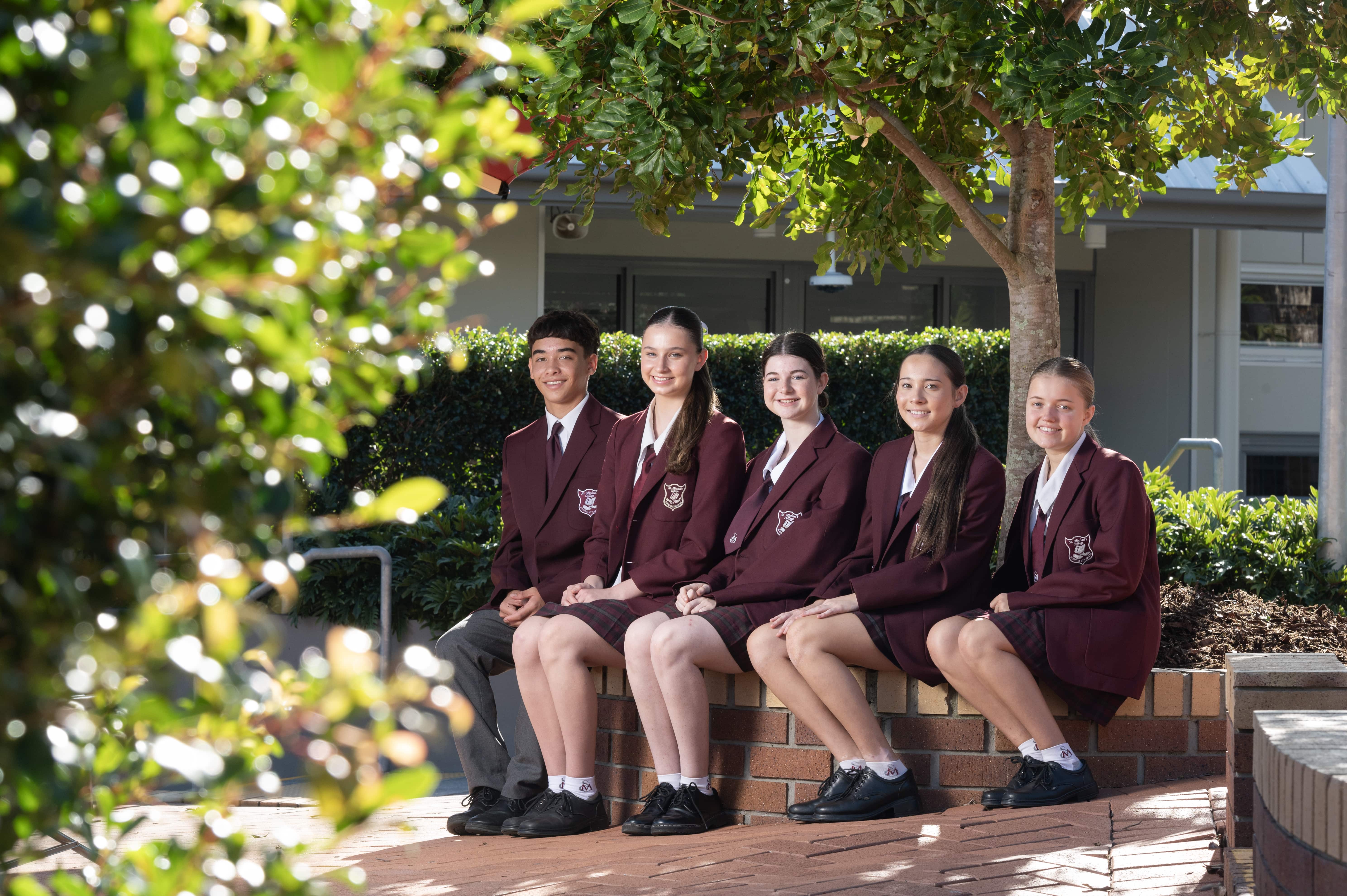 Students in school uniform seated together outdoors on a brick ledge, posing in a group within a landscaped school courtyard setting
