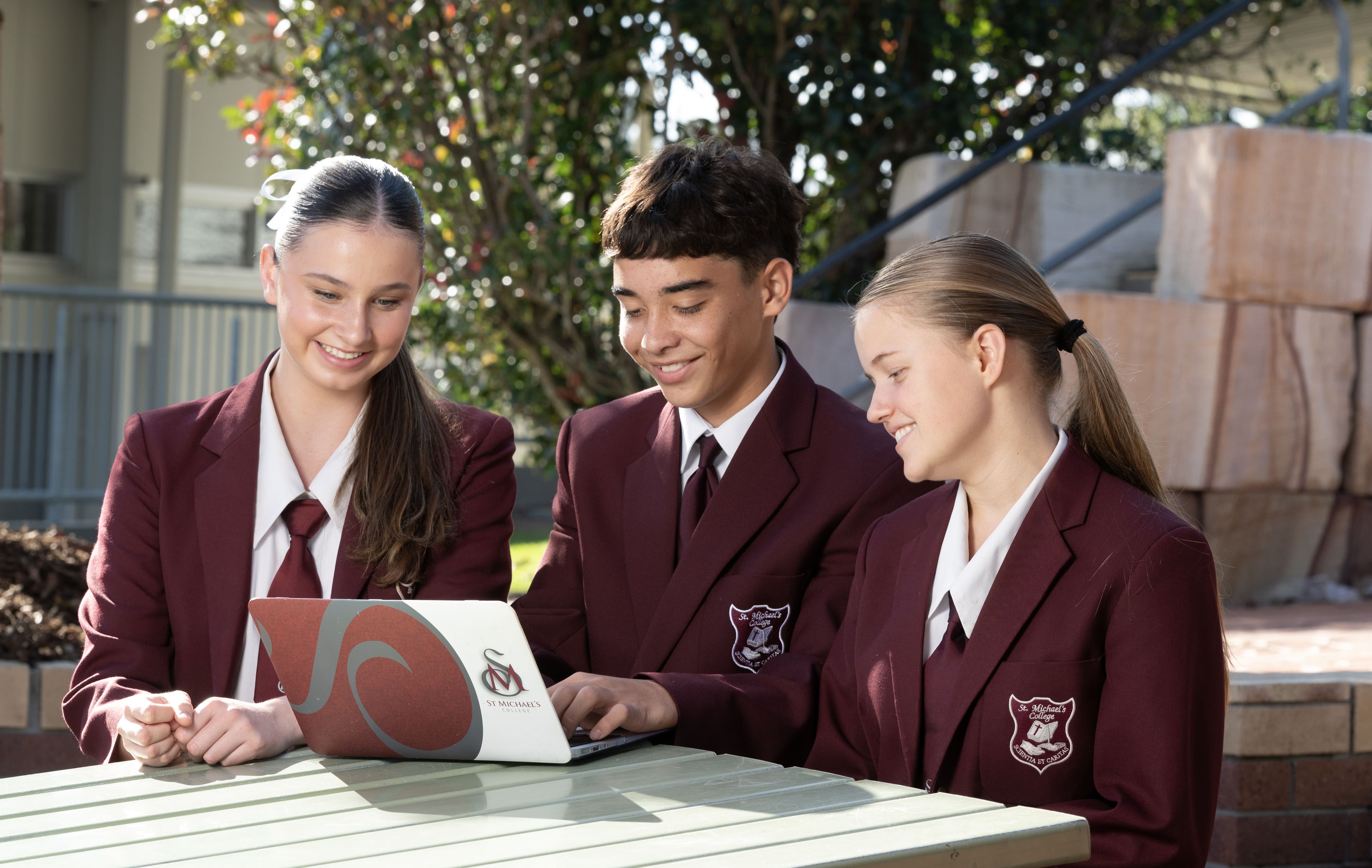 Students in school uniforms sitting outdoors at a table, using a laptop together and collaborating on a learning task