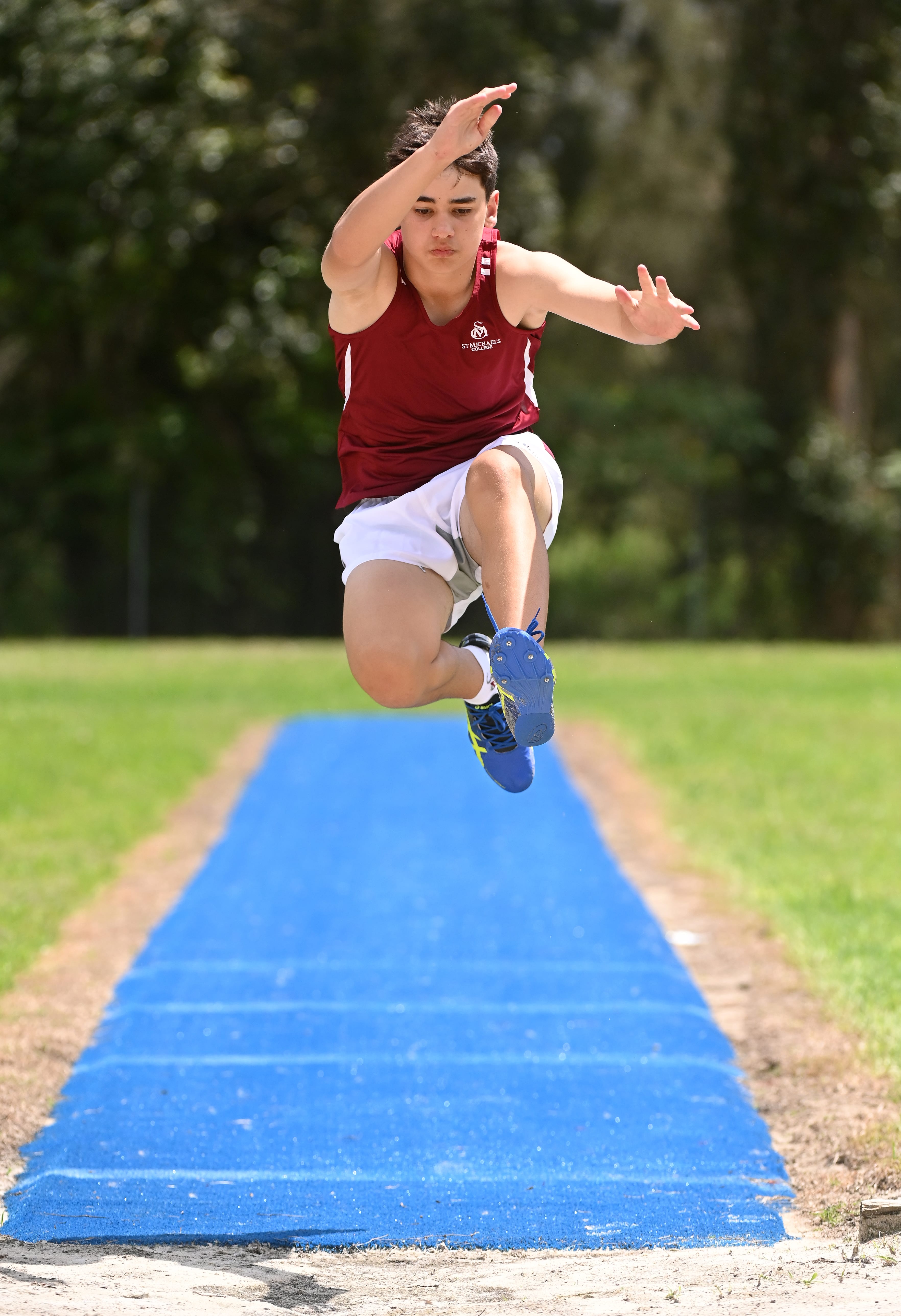 Student in sports uniform mid-air during a long jump, landing onto a blue track runway during an athletics event