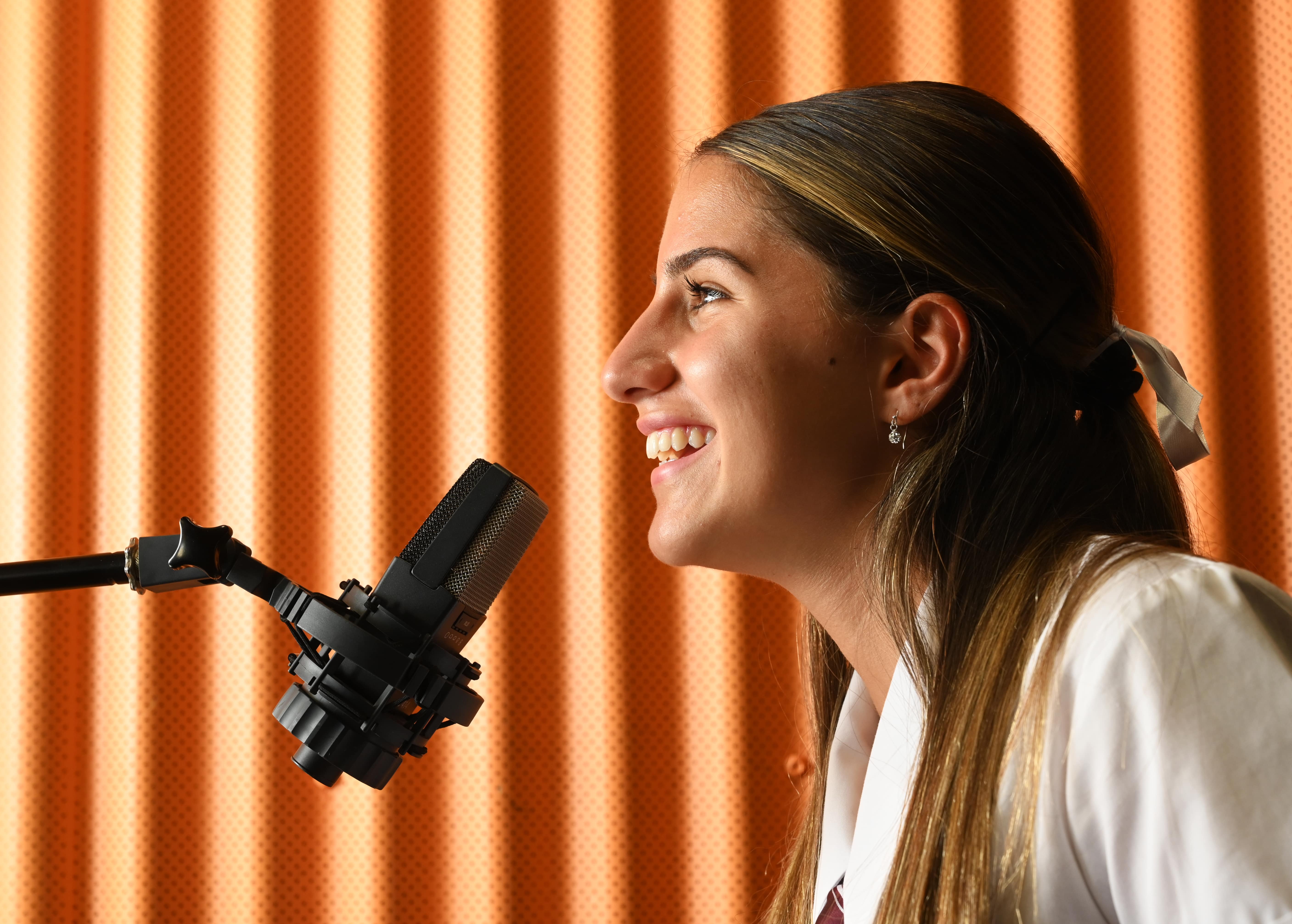 Student speaking into a studio microphone during a recording activity with acoustic backdrop in a school setting