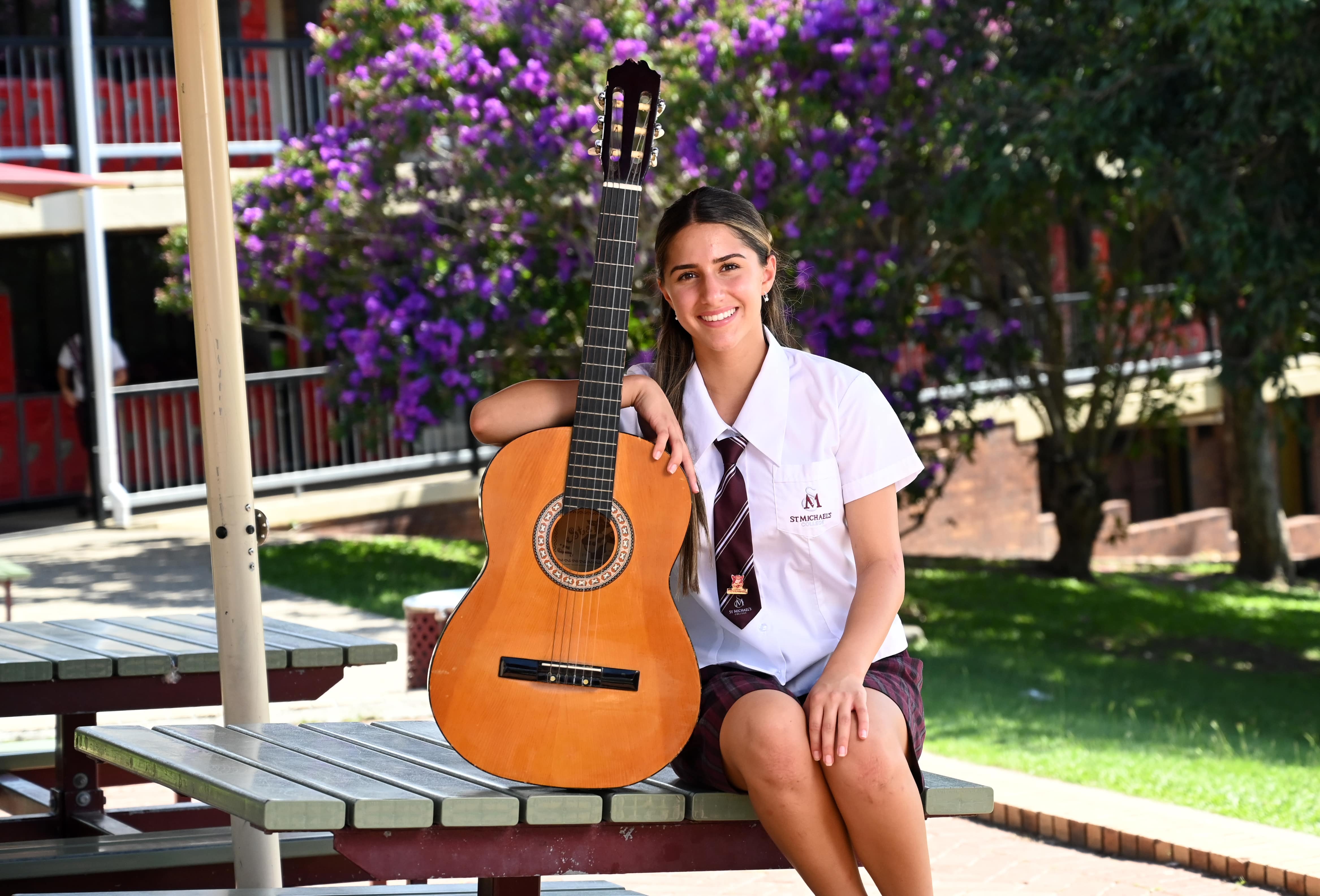 Student sitting outdoors holding a classical guitar in a school courtyard with flowering trees in the background