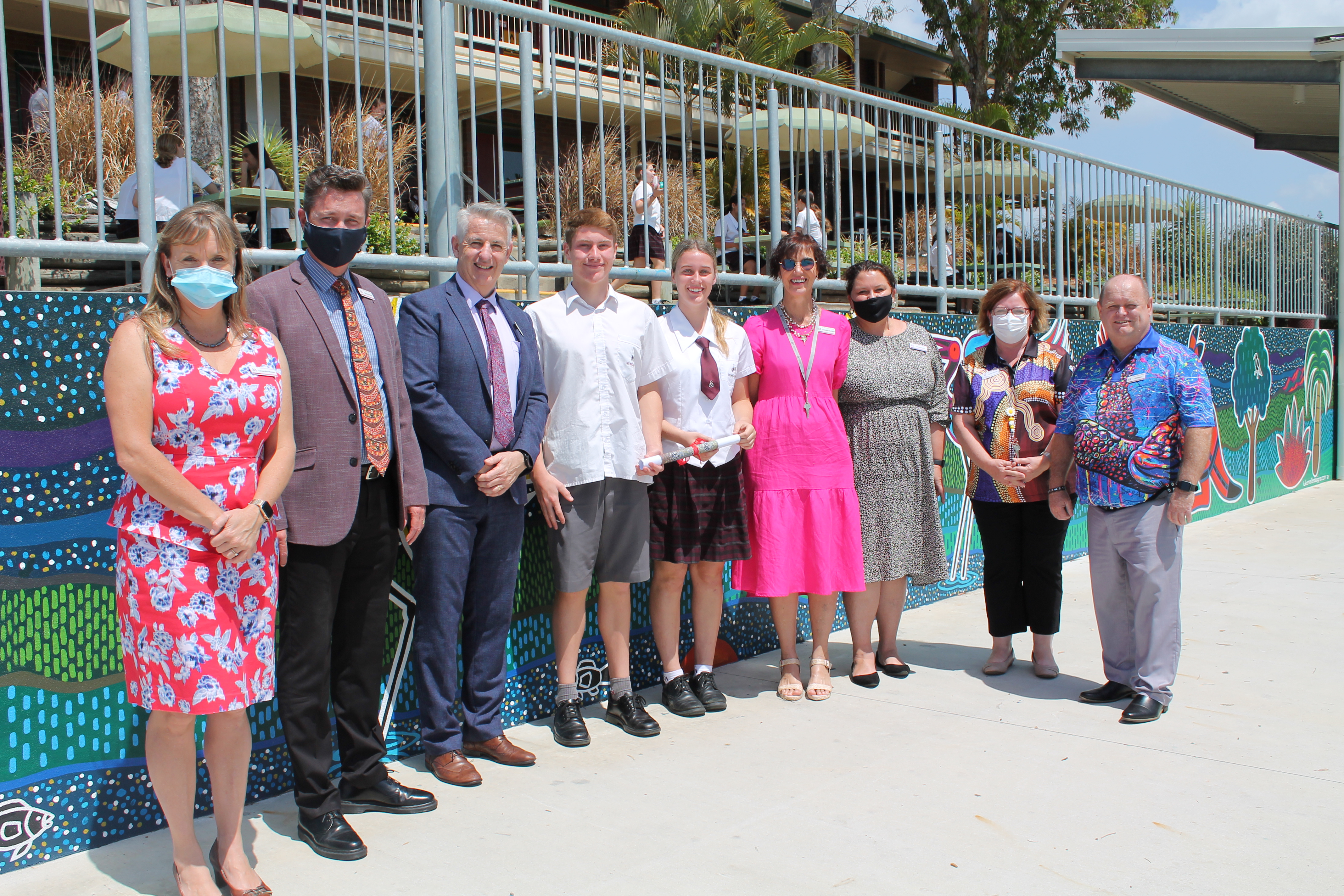 Staff, students and community members standing together outdoors beside a colourful mural on a school campus during an event