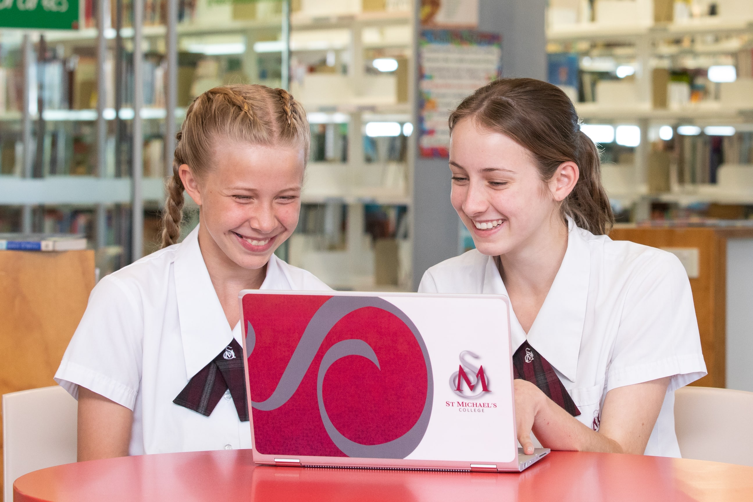 Students sitting together in a school library using a laptop, working collaboratively at a table with bookshelves in the background