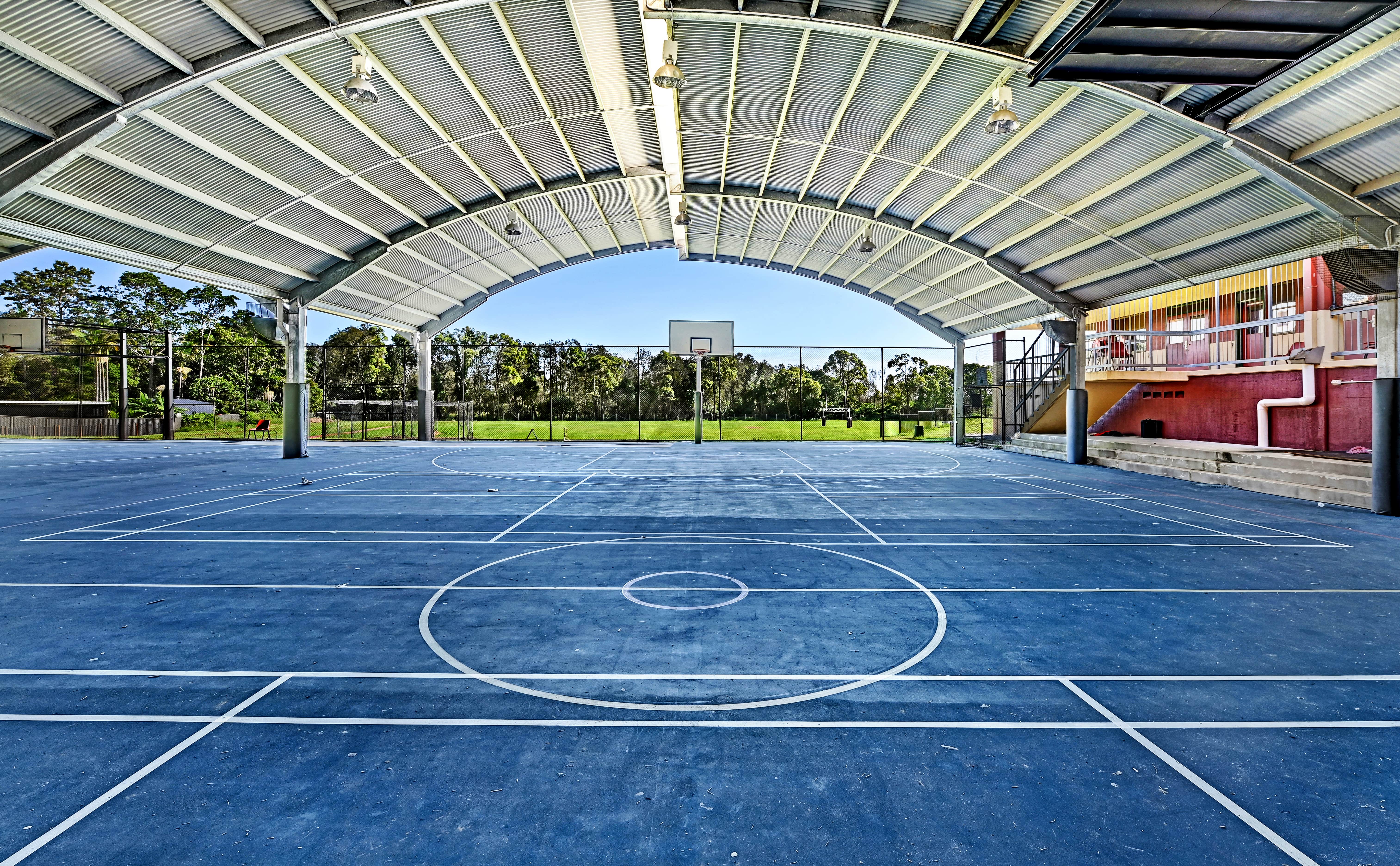 Covered outdoor basketball court with marked lines, hoop and open view to playing fields beyond